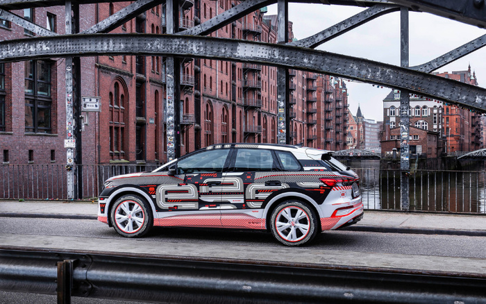 Modern electric SUV car with a futuristic black, white, and red patterned wrap featuring the text 'e-tron' parked on a street in front of historic brick warehouse buildings and a canal with metal bridge structures overhead, representing new cars and used cars in an urban setting.