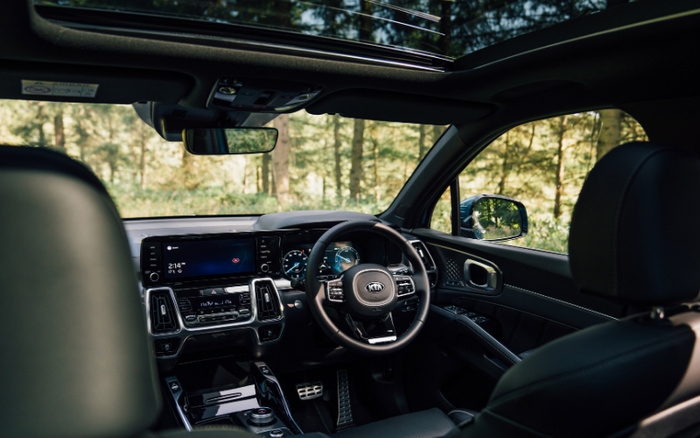 Interior view of a modern Kia car featuring a sleek black leather steering wheel with the Kia logo, a digital instrument cluster displaying a speedometer and other gauges, a central touchscreen showing 2:14 pm and a digital interface, climate control buttons, and a panoramic sunroof with a forest view outside. The image highlights the advanced and clean design of the car's cabin, relevant for new cars and used cars looking for a high-tech interior.