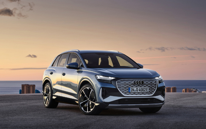 Blue Audi Q4 e-tron electric SUV parked on a rooftop with city buildings and sea in the background during sunset. The car features modern LED headlights, a large distinct grille with the Audi logo, and a European license plate reading 'IN Q 4002 E'. The image showcases the vehicle's sleek design and premium alloy wheels, ideal for highlighting both used cars and new cars in automotive sales or reviews.