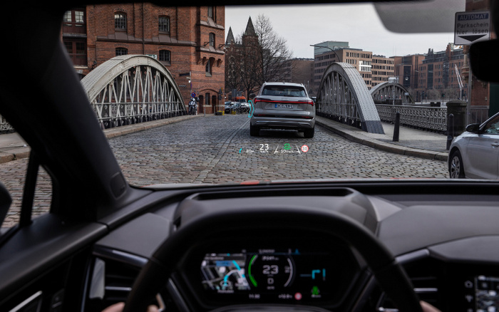 View from inside a modern car driving on a cobblestone street over an arched metal bridge in an urban area with historic brick buildings and contemporary apartments. The car ahead is a silver SUV with a visible license plate. The dashboard displays digital speed at 23 km/h, navigation directions, and other driving information on a heads-up display. A sign on the right reads 'AUTOMAT Parkschein hier lösen,' indicating a parking ticket machine. This image relates to used cars and new cars in city driving conditions.