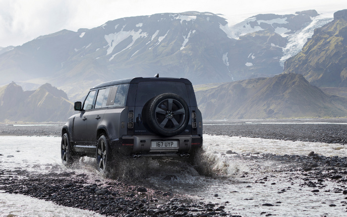 Black Land Rover Defender driving through shallow river water with rocky terrain, mountainous landscape with snowy peaks in the background, UK style number plate 167 UYP, showcasing rugged used cars and new cars in off-road conditions.