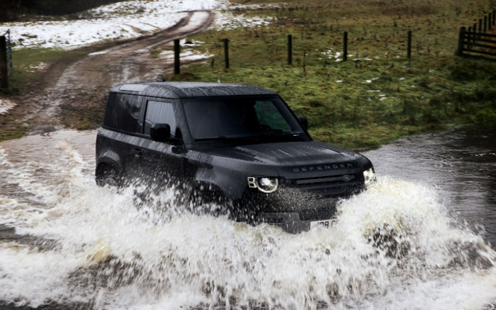 Black Land Rover Defender driving through a large water puddle on a muddy off-road track with grassy fields and patches of snow in the background, illustrating capability and ruggedness relevant to used cars and new cars.