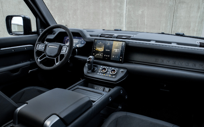 Interior of a Land Rover Defender showing the modern dashboard and steering wheel with digital display and central touchscreen featuring media and climate controls. The black interior highlights the detailed stitching and high-quality materials, ideal for used cars and new cars buyers seeking a premium vehicle experience.