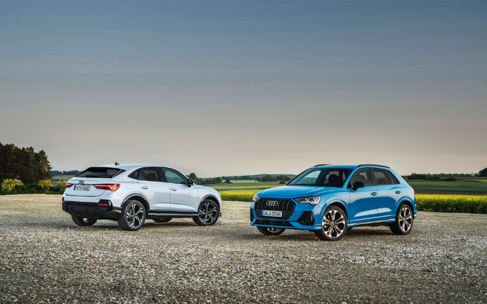 Two Audi SUVs parked on a gravel area with a countryside background under a clear sky. One is a white Audi Q3 Sportback viewed from the rear left side with license plate IN-Q320E, and the other is a blue Audi Q3 viewed from the front right side with license plate IN-Q320E. Both vehicles are new cars showcased outdoors, suitable for buyers interested in used cars or new cars.