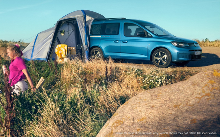 Blue Volkswagen camper van parked on a grassy, rural campsite next to a pitched grey and blue tent. A young girl in a pink shirt and white shorts walks nearby holding a bottle. The scene captures a sunny day with dry grass and wildflowers. Text in the bottom right corner reads 'Image for illustration purposes only. Vehicle shown may not reflect UK specification.' This image is suitable for illustrating used cars or new cars in outdoor leisure settings.