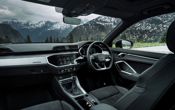 Interior of a modern Audi car showing the driver's seat, steering wheel with Audi logo, and dashboard featuring a digital display and control panel against a backdrop of snow-capped mountains and pine trees visible through the windows. The image highlights the sleek design and technology of the car, suitable for used cars and new cars marketing.