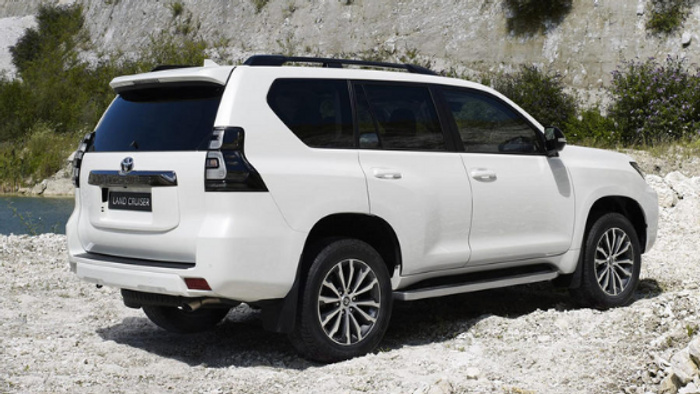 White Toyota Land Cruiser SUV parked on a rocky, gravel surface by a body of water with a rocky cliff and sparse greenery in the background, showcasing the vehicle's side and rear view, representing available used cars and new cars.