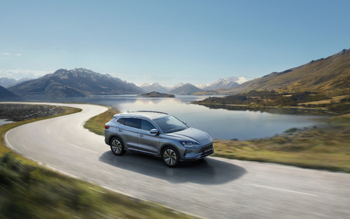 Silver SUV driving on winding road by a serene lake with mountains in the background, showcasing a modern new car model ideal for those interested in buying new cars or used cars in scenic locations.