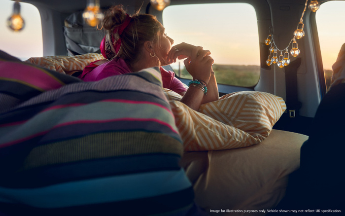 Woman lying down inside a vehicle at sunset, resting her head on a striped cushion and looking out the window. The vehicle interior is decorated with multiple cushions and hanging string lights. Text at the bottom right corner reads: 'Image for illustration purposes only. Vehicle shown may not reflect UK specification.' This image evokes a relaxed and cozy atmosphere, suitable for illustrating concepts related to used cars and new cars.