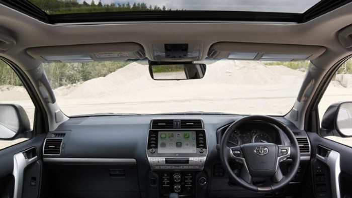 Interior view of a Toyota car from the driver's perspective showing the steering wheel with Toyota logo, dashboard with digital touchscreen display, climate controls, side mirrors, and panoramic sunroof. This image highlights the modern features and spacious design typical in both used cars and new cars.