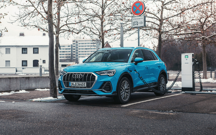 Blue Audi electric vehicle charging at a public electric charging station in a parking spot marked by a sign prohibiting parking except for electric vehicles, with the sign showing a car and a charging plug symbol. The car has a license plate reading IN Q3440E, and the scene includes leafless trees and buildings in the background in winter conditions with some snow on the ground. This image is relevant for used cars and new cars, particularly electric or hybrid models.