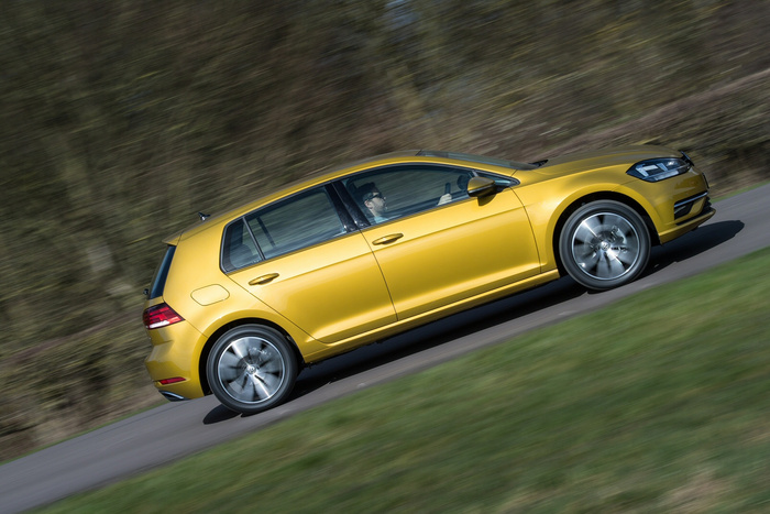 Yellow Volkswagen Golf hatchback in motion on a paved road with blurred trees and grass in the background, driven by a person wearing glasses. The image represents used cars and new cars.