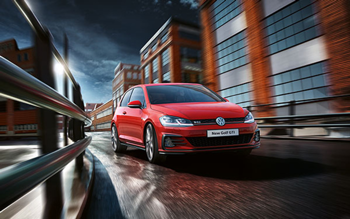 Red Volkswagen New Golf GTI car driving on a wet urban road with modern brick and glass buildings in the background at dusk, showcasing a sporty used car or new car option.