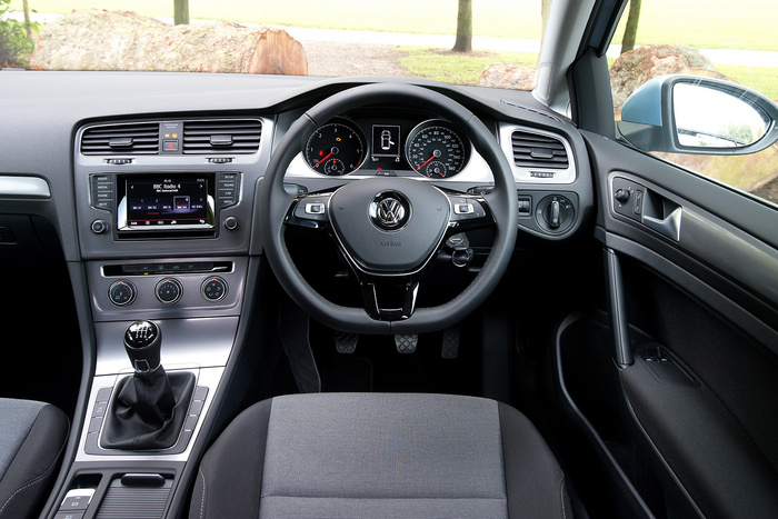 Interior view of a Volkswagen car showing the steering wheel with the VW logo, a manual gear stick, and dashboard. The dashboard display is tuned to BBC Radio 4 on DAB with options for Radio, Media, Phone, Mute, Setup, Sound, Car and Menu visible on buttons. The speedometer and tachometer dials are clearly visible, along with air conditioning controls and a passenger airbag indicator. The car features black and grey fabric seats and the outside view shows a park with grass and trees. Ideal representation of used cars and new cars with modern interior features.