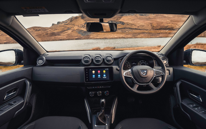 Interior view of a Dacia car showing the dashboard, steering wheel with Dacia logo, touchscreen infotainment system, air vents, and gear shift. The car is right-hand drive, with a scenic landscape of mountains and a lake visible through the windshield and side windows. Suitable for showcasing used cars and new cars.