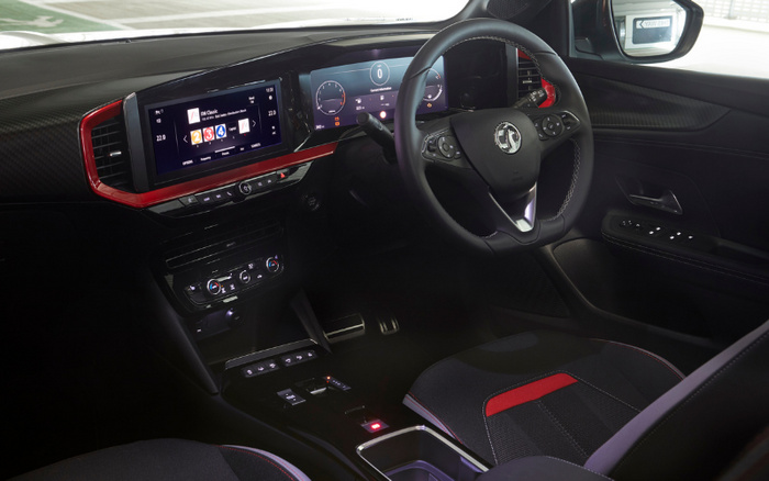 Interior view of a modern car featuring a digital driver's dashboard and central touchscreen display showing a music playlist, a black steering wheel with a Vauxhall logo, red trim accents on the dashboard, climate controls below the screen, and fabric seats with red stitching and detailing, highlighting features commonly found in both used cars and new cars.