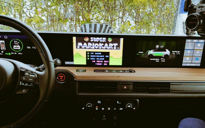 Dashboard and interior of a modern electric vehicle featuring a wide digital display panel. The display shows a game screen with the title 'Super Mario Kart' and options for 1P Game, Mario Kart GP, 50cc Class, and selecting 'Yes' or 'No'. Next to the game screen, the display indicates the car's charge status at 91%, remaining time of 17 minutes, and a graphic of the car in charging mode. The steering wheel and control buttons are visible, highlighting modern car technology in a new car, suitable for used cars seeking advanced digital features.