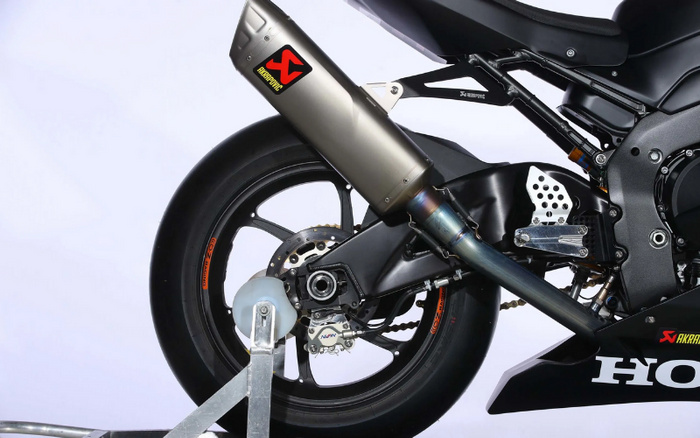 Close-up of the rear wheel and exhaust system of a black Honda motorcycle. The exhaust has a silver Akrapovic muffler with the Akrapovic logo in black and red on a yellow background. The wheel rim has an orange OZ Racing decal. The motorcycle is secured on a metal rear stand with a white roller. The image highlights motorcycle components, relevant to enthusiasts of used cars and new cars looking for performance vehicle options.