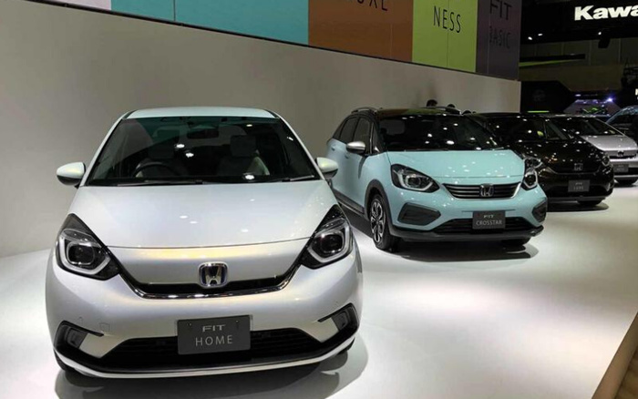 Display of new cars at a showroom featuring a white Honda Fit Home and a light blue Honda Fit Crosstar in the foreground, with additional cars lined up behind them. The vehicles are showcased on a well-lit platform with branding and text panels overhead, highlighting models available for potential buyers interested in used cars and new cars.