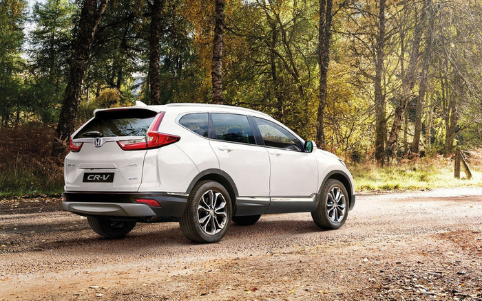White Honda CR-V hybrid parked on a dirt road surrounded by trees in autumn. The rear and side view of the car show distinct taillights and alloy wheels, with badges indicating 'e:HEV' hybrid technology. The setting suggests a spacious and reliable SUV ideal for both new cars and used cars buyers seeking versatility and eco-friendly features.