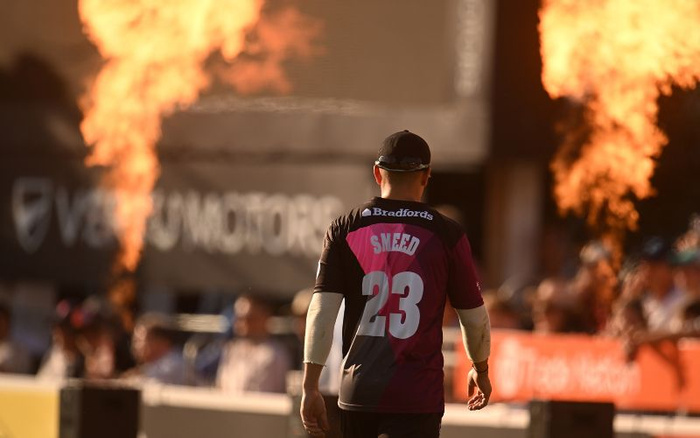 Cricket player wearing a black and maroon jersey with the name 'Smeed' and number 23 on the back walking away from the camera. The jersey features a 'Bradfords' logo across the shoulders. In the background, there are flaming bursts on both sides and a blurred crowd behind a barrier with a partially visible sign that reads 'VE... MOTORS', suggesting a car dealership or sponsor likely related to new cars and used cars.