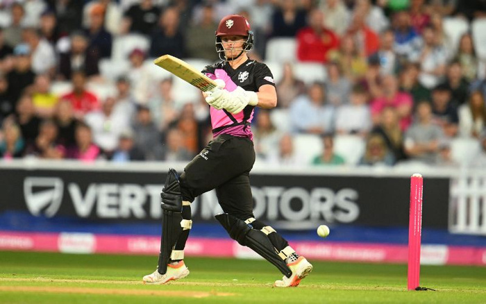 Cricket player in black and pink kit swinging a bat towards the ball during a match, with an advertising board in the background displaying the text 'VERTEX MOTORS' and 'used cars new cars' partially visible.