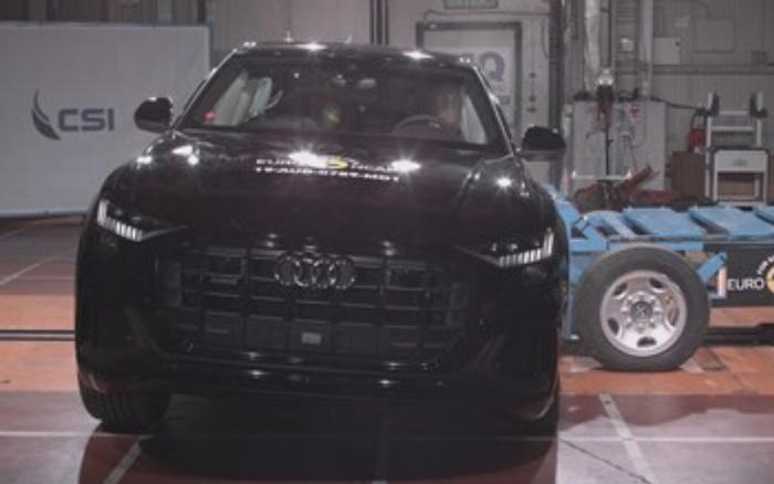 Front view of a black Audi car inside an industrial testing facility with a blue and white equipment vehicle or testing rig beside it. The car has the Audi logo prominently displayed on its black grille. The headlamps are turned on, showing distinct LED lines. In the background on the left, there is a white banner with the text 'CSI' and a stylised wing logo. The setting appears to be indoors with visible tools and machinery. This image relates to new cars and used cars in an automotive testing or showroom environment.