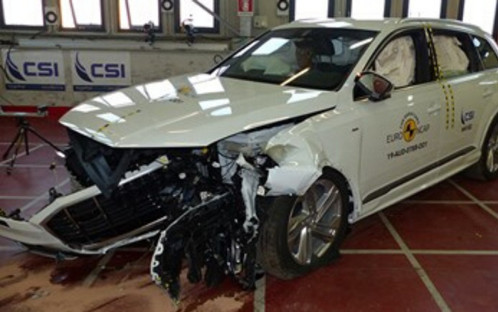 White car with severe front-end crash damage involved in a Euro NCAP crash test, showing crushed front bumper and exposed engine components, with the Euro NCAP Thatcham Research logo on the side. The car is parked indoors on a tiled floor with CSI banners in the background. This image relates to safety testing relevant for used cars and new cars.