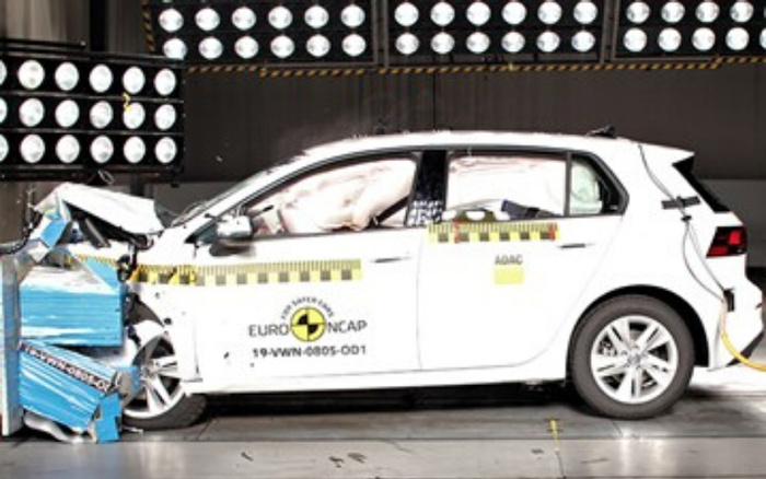 White hatchback car undergoing a frontal crash test with airbags deployed, inside a crash test facility with multiple circular lights on the wall. The car has Euro NCAP logo and identification code 19-WVW-4085-001 on the side, along with ADAC labels. The vehicle is connected to monitoring equipment, illustrating a safety test for used cars and new cars.