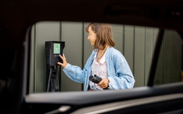 Young woman charging an electric car at a residential EV charging station, holding the charging plug and pressing a button on the charger, viewed through the open car window. The charging station display shows charging information. Ideal image for used cars and new cars electric vehicle promotion.