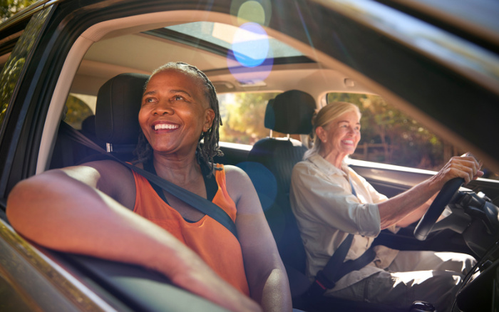 Two smiling women in a car, one driving and the other in the passenger seat, enjoying a sunny day. Image suitable for used cars and new cars advertisement.