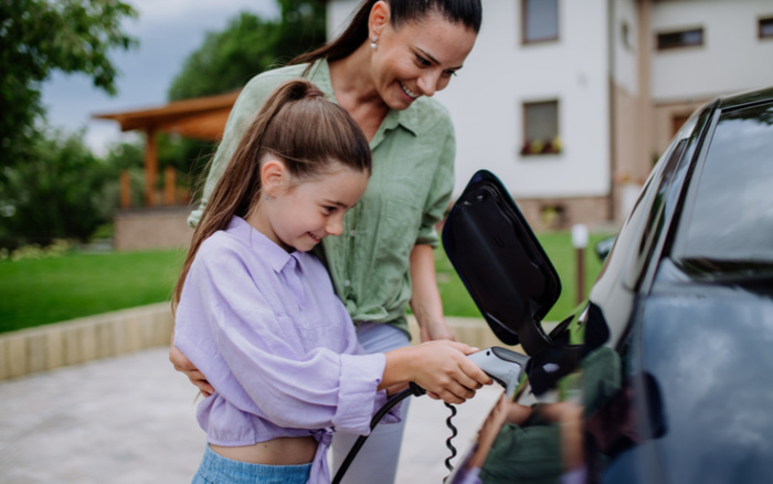 Mother and daughter smiling while charging an electric car at home, demonstrating eco-friendly used cars and new cars technology, with a suburban house and garden in the background.