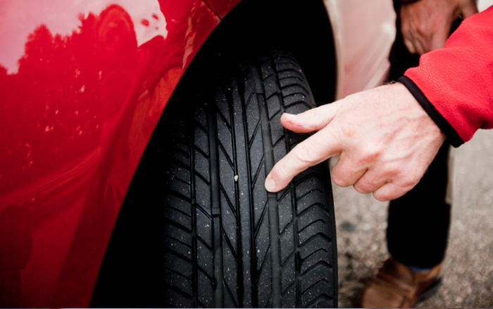 Close-up of a person pointing at a small stone lodged in the tread of a black car tyre next to a red car body, highlighting tyre condition important for safety when buying used cars or new cars.