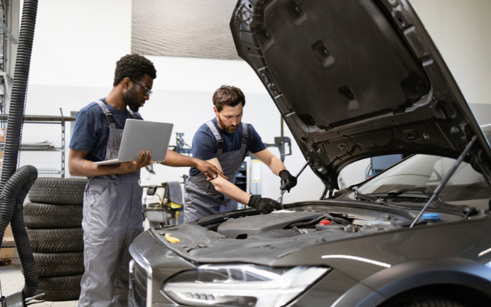 Two mechanics in a workshop inspecting and repairing a car with its hood open; one mechanic is using a laptop while the other is working on the engine with a tool; stacks of tires are visible in the background, representing maintenance and service for both used cars and new cars.