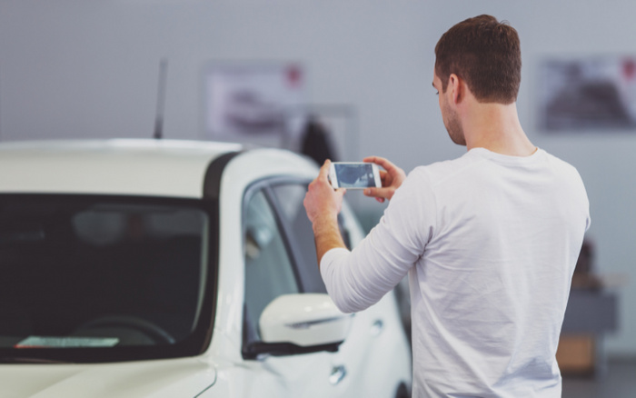 Man in white long-sleeve shirt taking a photo of a white car using a smartphone inside a car dealership showroom, representing interest in used cars or new cars.