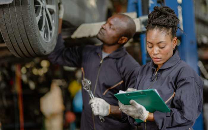 Two mechanics in a garage working on a car lifted on a hydraulic lift, one inspecting the underside and wheel of the car while holding a wrench, the other checking notes on a green clipboard, both wearing dark work uniforms and gloves, representing car servicing for both used cars and new cars.