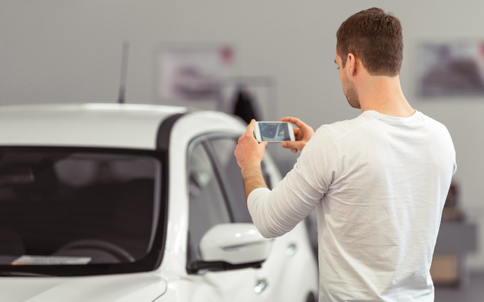 Man in white long-sleeve shirt taking a photo of a white car inside a showroom, likely capturing details for buying or selling used cars or new cars.