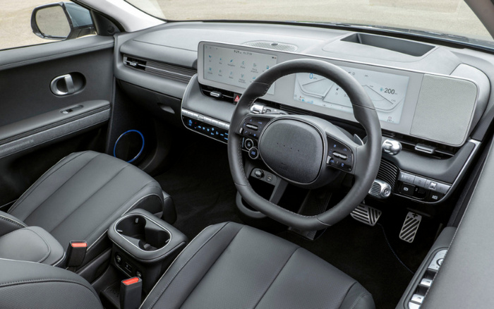 Interior view of a modern car with right-hand drive, featuring black leather seats, a large digital dashboard display showing speed and settings, a digital infotainment screen with icons such as Bluetooth, media, and phone, a multifunction steering wheel with control buttons, and a central console with cup holders and seatbelt buckles. The design is sleek and contemporary, suitable for new cars and used cars buyers.