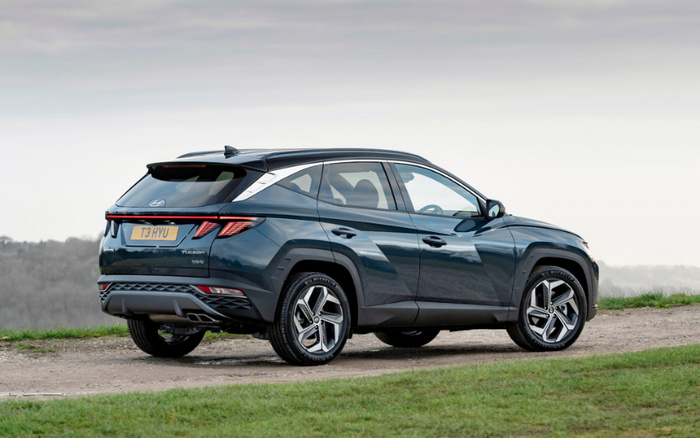 Rear and side view of a dark blue Hyundai Tucson hybrid SUV parked on a gravel path surrounded by grass with a cloudy sky background. The vehicle has UK registration plate T3 HYU. This image is suitable for advertising used cars and new cars, showcasing a modern hybrid SUV model.