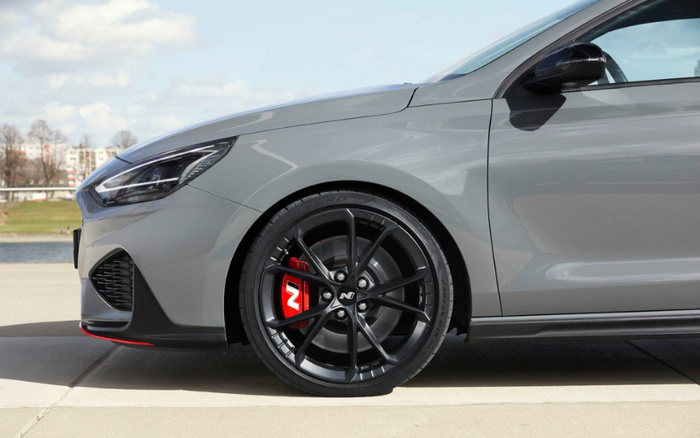 Close-up side view of a sleek grey car focusing on the front wheel with black alloy rims and a red brake caliper featuring a white 'N' logo, parked outdoors on a concrete surface with a blurred background of grass, water, trees, and buildings, suitable for used cars and new cars.