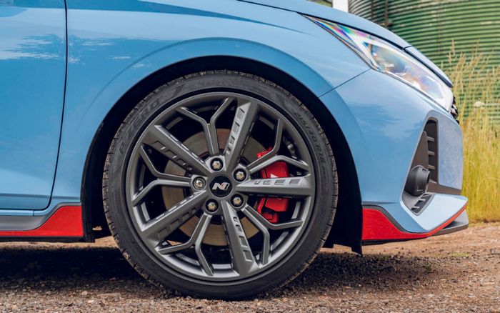 Close-up of a blue new car's front wheel and side panel, featuring a grey stylish alloy rim with an 'N' logo at the centre and a red brake caliper. The car has modern LED headlights and a sporty front bumper with red accents. The image highlights the vehicle's design details, suitable for used cars and new cars listings.