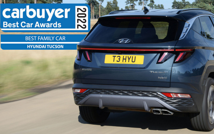 Rear view of a blue Hyundai Tucson hybrid driving on a country road with a yellow license plate reading T3 HYU. The image includes a blue and white banner on the top left corner that says 'carbuyer Best Car Awards 2022', awarding the Hyundai Tucson as the Best Family Car. The photo highlights used cars and new cars as relevant keywords.