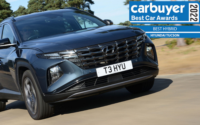 2022 Carbuyer Best Car Awards banner showing the Best Hybrid award won by the Hyundai Tucson, with a close-up image of a dark blue Hyundai Tucson car driving on a road. The car's front grille, headlights, and license plate reading 'T3 HYU' are clearly visible. Keywords related to used cars and new cars.