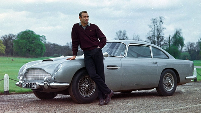 Classic silver Aston Martin DB5 parked on a gravel driveway with a man in a burgundy sweater and dark trousers leaning against the front wing. The car has a vintage design featuring wire spoke wheels and a front registration plate BMT 216A. The scene is set in a green open area with trees in the background. Image related to used cars and new cars, showcasing a timeless luxury vehicle.