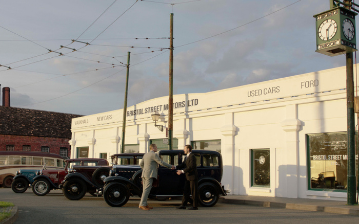 Two men shaking hands beside a vintage black car in front of a classic white car dealership building with signage reading 'VAUXHALL NEW CARS', 'BRISTOL STREET MOTORS LTD', 'USED CARS', and 'FORD'. Several classic cars are parked outside the dealership, which has a large clock overhead showing the time as 9:30. The scene captures a nostalgic atmosphere related to used cars and new cars at Bristol Street Motors.