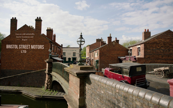 Street scene with historic brick buildings and a vintage red car parked near a bridge over a canal. Sign on a building reads 'Cars are sold universally, but for Courteous and Expert BRISTOL STREET MOTORS LTD'. Another building in the background has a sign for 'BOTTLE & GLASS INN'. The image evokes a classic car dealership setting, relevant to used cars and new cars.