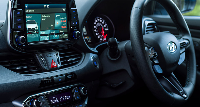 Interior dashboard of a Hyundai car showing the central display screen with performance and g-force data, including turbo, torque, and power meters. The screen shows a white car image with 'N mode' at the top left and time 11:30 at the top centre. Controls for radio, media, and seek track are visible on the side of the display. Below the screen is the hazard light button and air vents, with climate control settings below showing air conditioning at 22°C. The steering wheel features Hyundai's logo and various control buttons. This modern car interior is relevant for both used cars and new cars.