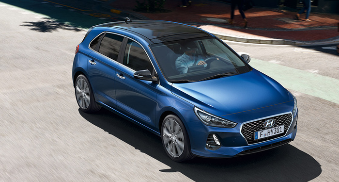 Blue Hyundai hatchback driving on a city street with a driver visible inside, featuring modern LED headlights and a black roof. The car has a European license plate reading 'F HY301'. Ideal for used cars and new cars buyers looking for a compact, stylish vehicle.