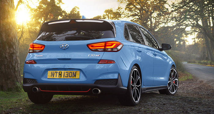 Rear view of a blue Hyundai i30 N hatchback car parked on a forest road during sunset with illuminated rear lights and a yellow UK license plate reading NTH130N, showcasing a sporty exhaust system and alloy wheels, representing both used cars and new cars.