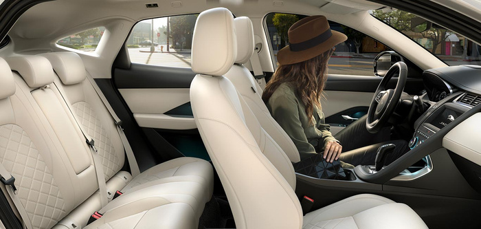 Interior view of a luxury car with cream quilted leather seats showing front and rear passenger areas, a woman wearing a brown hat and green jacket sitting in the driver's seat, visible Jaguar logo on the infotainment screen, modern dashboard and steering wheel, with a street scene visible through the windows, highlighting the comfort and style suitable for used cars and new cars buyers.